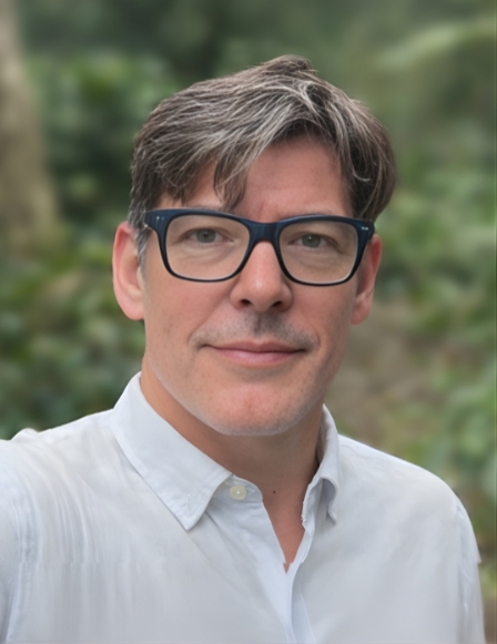 A member of the Leadership Team with short brown and gray hair, wearing black glasses and a white button-up shirt, stands outdoors with greenery in the blurred background.