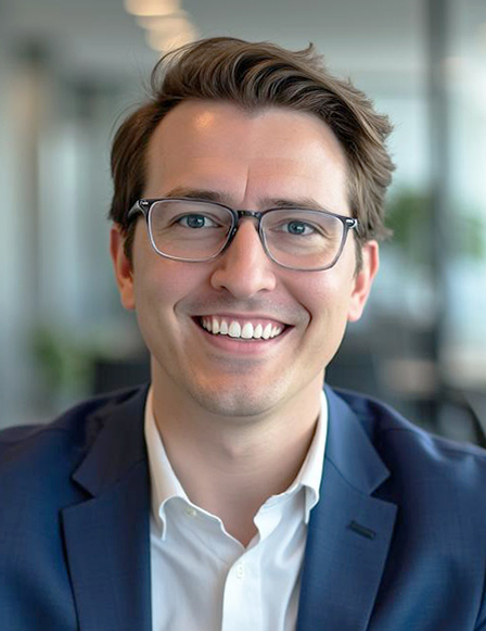 A member of the Leadership Team, this man with brown hair and glasses, wearing a navy suit and white shirt, smiles at the camera in an office setting.